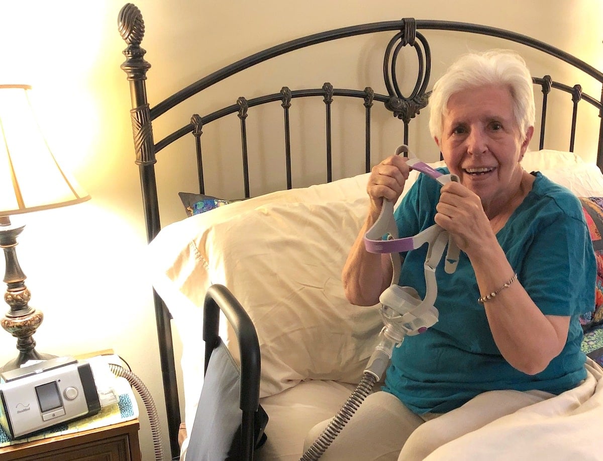 A woman sits on the side of her bed holding an NIV mask.